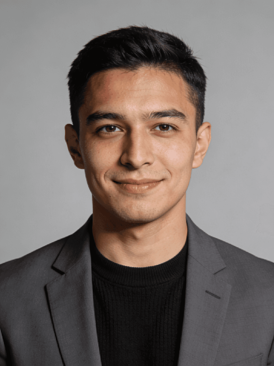 A young man with short dark hair, wearing a gray blazer and black shirt, smiling against a plain gray background.