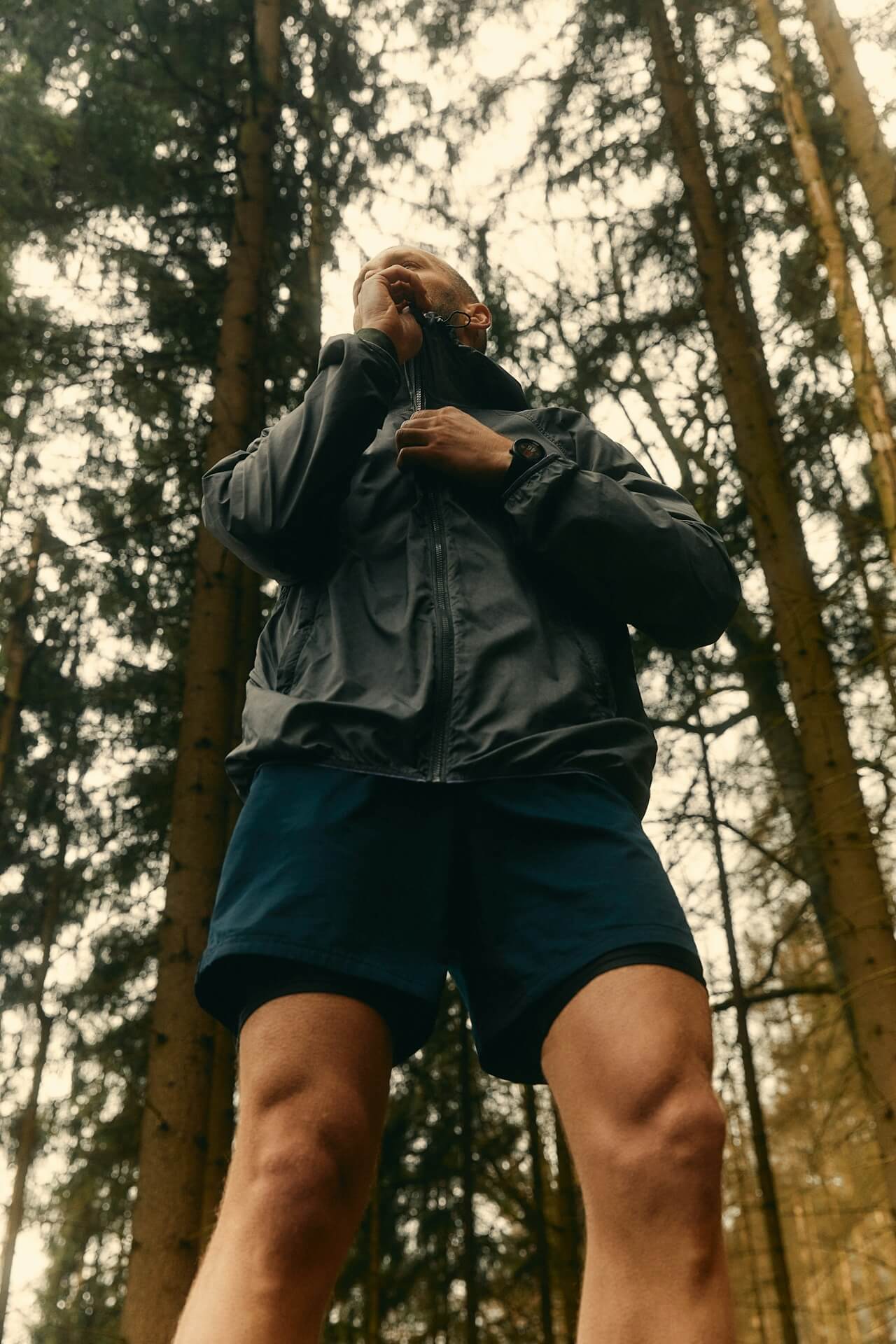 A person in a gray jacket zips up, standing confidently in a dense forest. Tall trees surround them, creating a serene and adventurous mood.