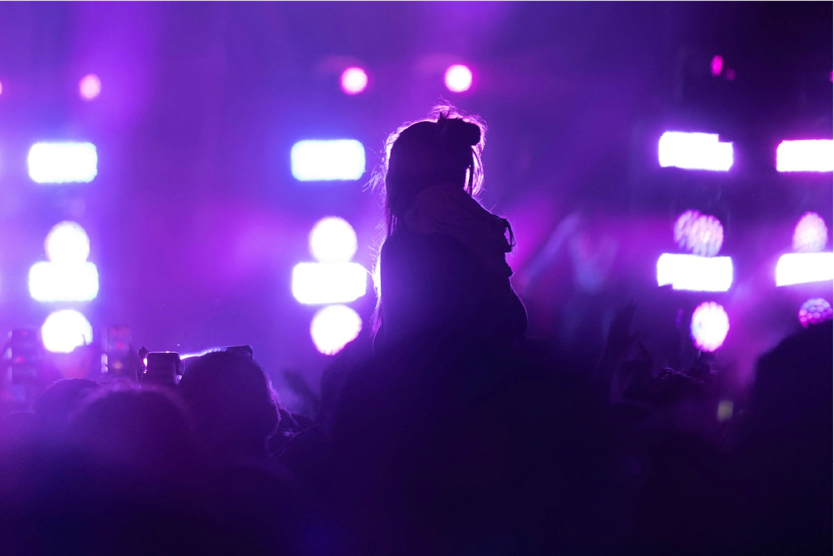 Silhouette of a person in a crowd, backlit by bright purple and white stage lights.