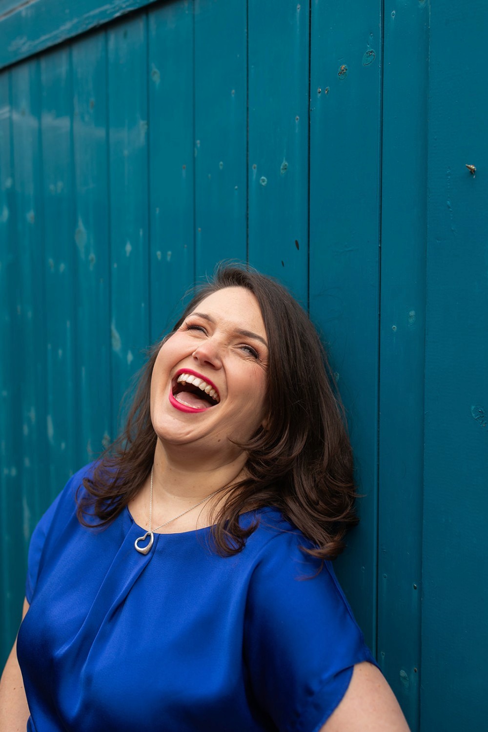 Lucy with her head thrown back in laughter, in front of a bright blue wall.