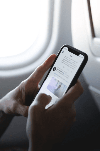 A person holding a smartphone and viewing a social media post while seated near an airplane window. The image background is light gray with a pattern of small, evenly spaced gray dots on the left side.