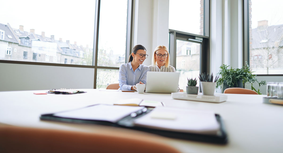 Two coworkers smiling while sharing a laptop screen in a bright conference room with a planner
