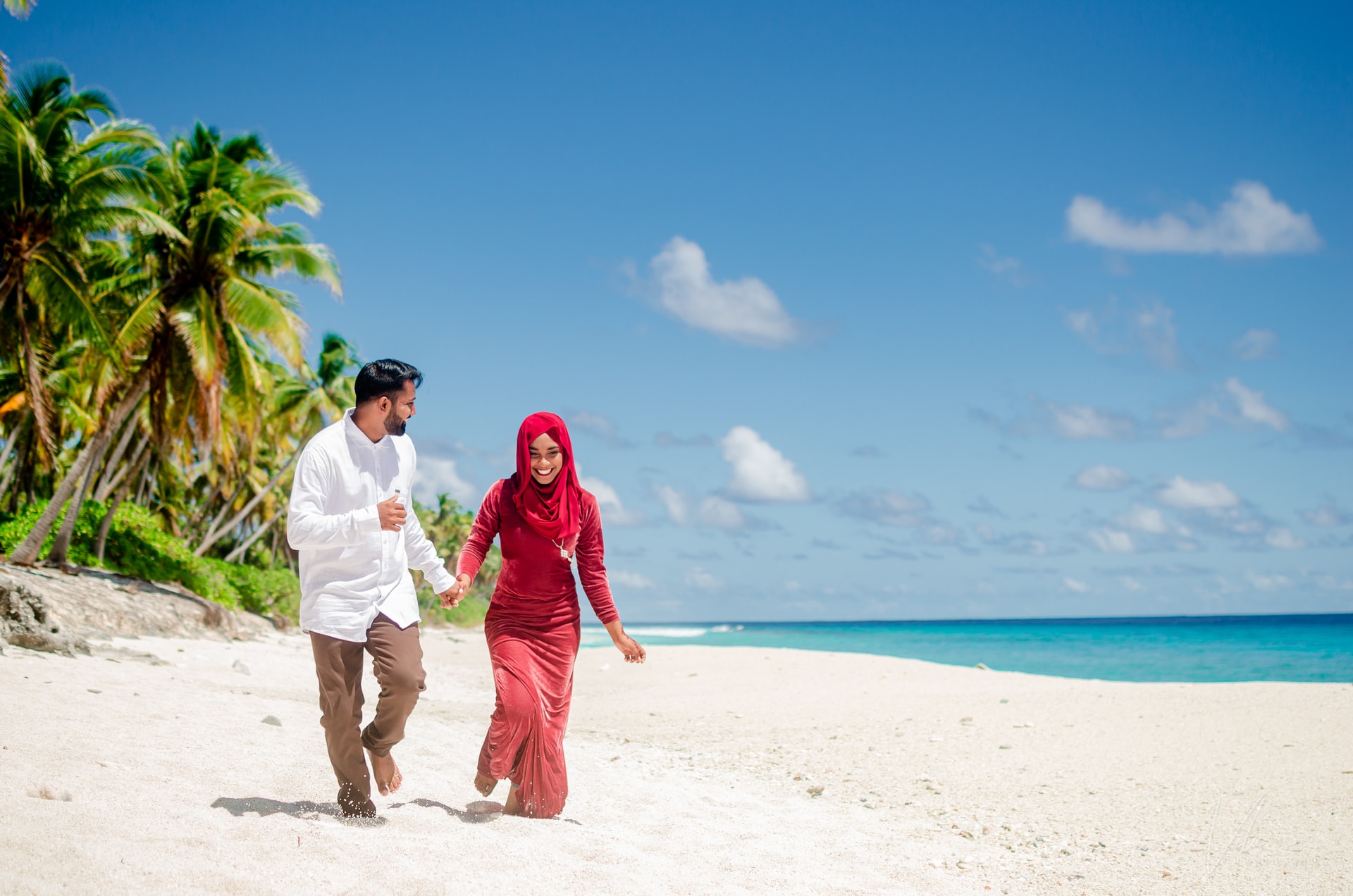 A couple walks hand-in-hand on a beach, surrounded by palm trees and a clear blue sky.