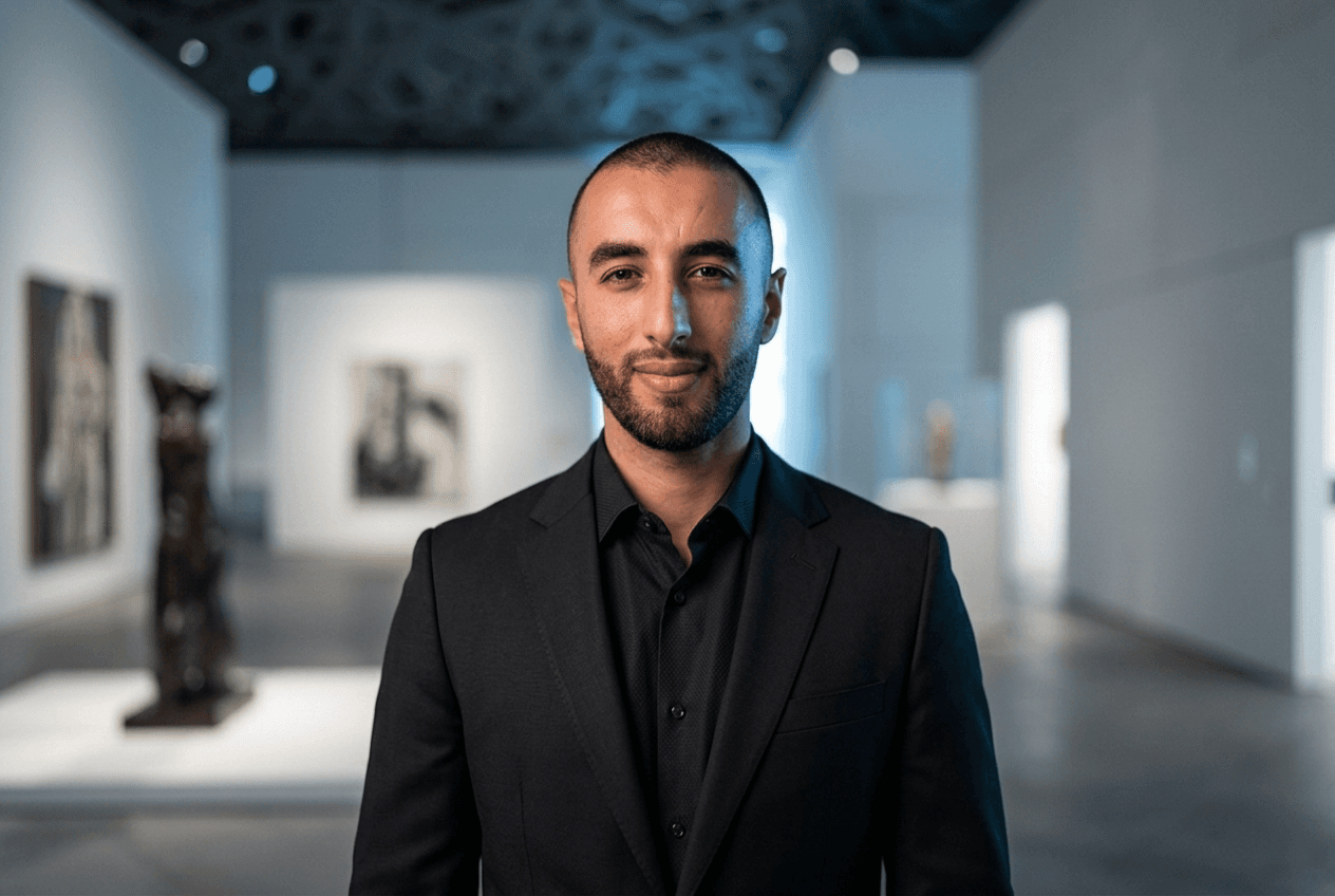 Man in black suit standing in modern art gallery with sculptures and paintings in softly lit background.