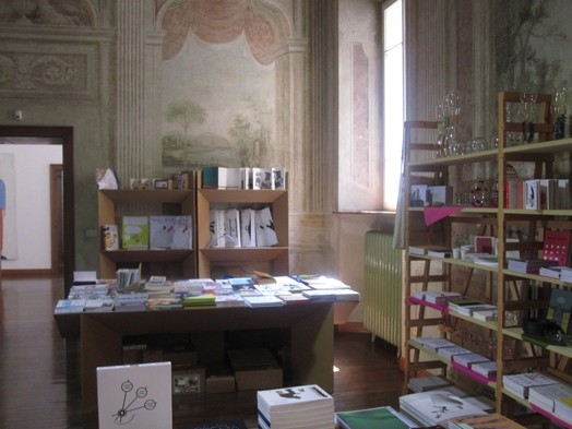 A photo of a library with books on a table and also stacked on shelfs.