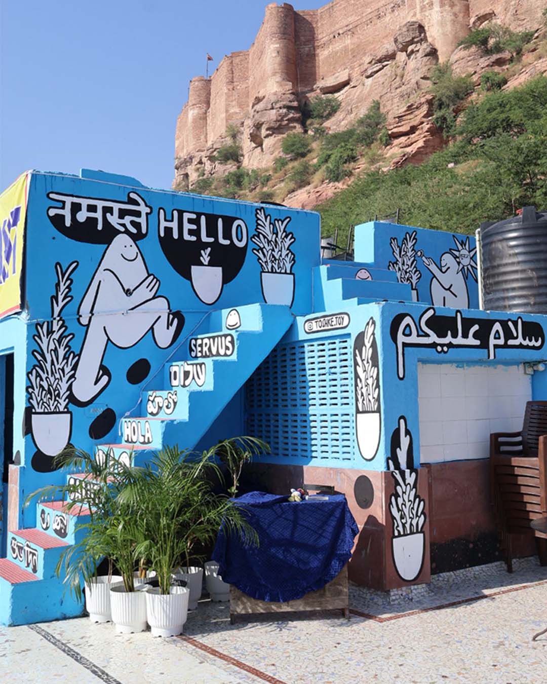 Mural painting with joyful cartoon characters and the word ‘Hello, नमस्ते, سلام عليكم’ painted in black-and-white on a terrace of a blue building near Jodhpur Fort.