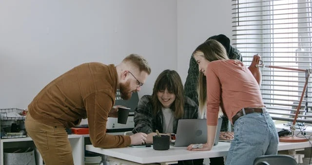 Três pessoas em volta de uma mesa com laptops num escritório claro, uma delas de pé apontando para a tela