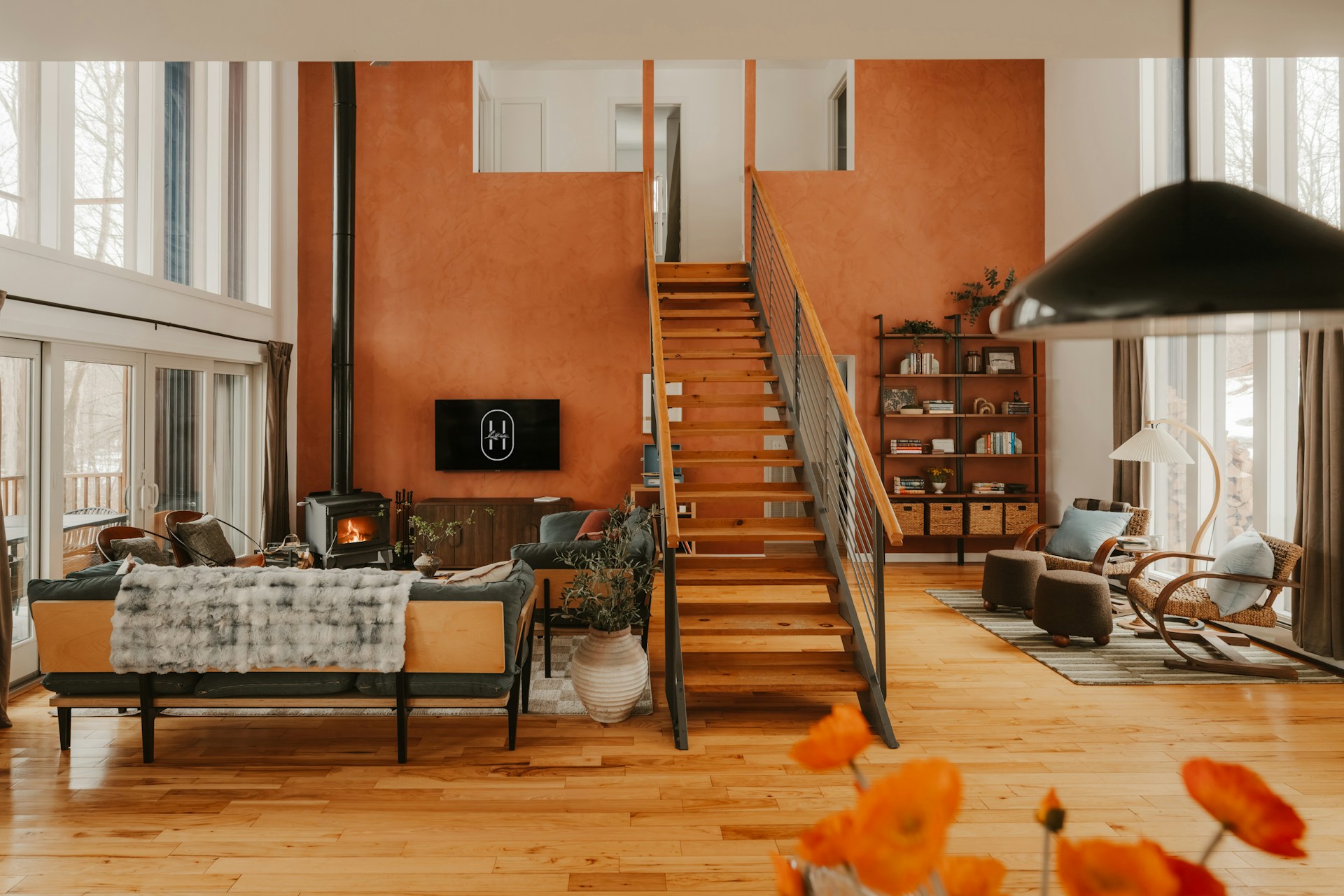 Cozy living room with a rust wall, central wooden stairs, wood-burning stove, sofa, and floor-to-ceiling windows.
