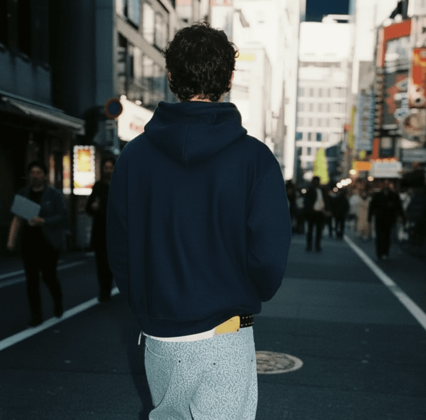 Back view of person with curly dark hair wearing a navy hoodie and light gray pants walking down a busy urban street in Japan with pedestrians and buildings visible