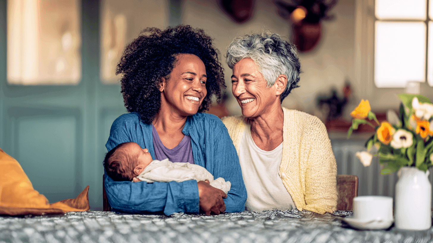 A joyful young woman and an older woman smile together, holding a baby in a cozy indoor setting.