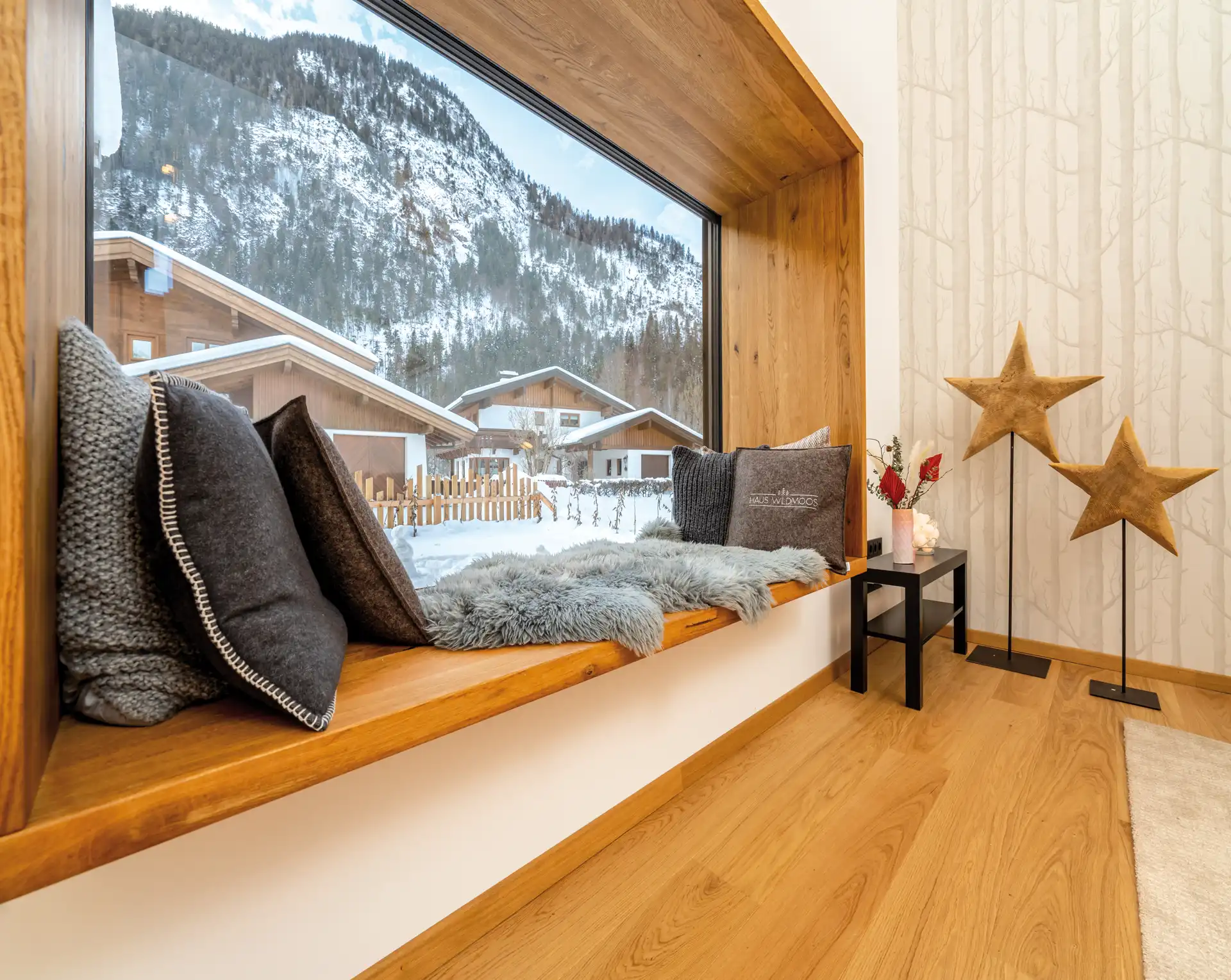 Holzbalkon mit Kissen, Fellen und Blick auf verschneite Alpenhäuser; daneben zwei Sternenständer.