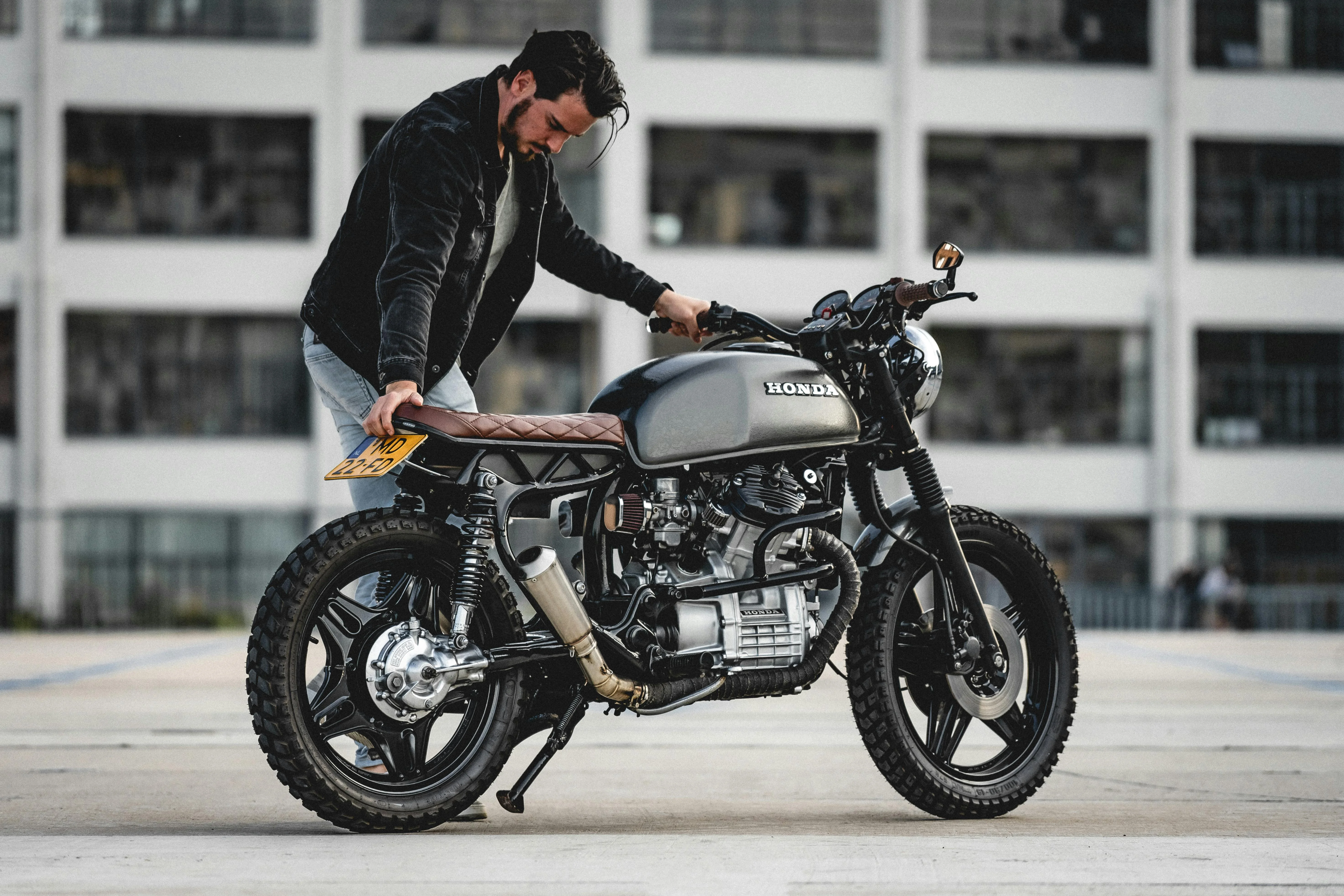 Moto Honda customisée dans un style café racer, mise en scène avec son pilote dans un décor urbain. Selle en cuir marron, pneus crantés, cadre retravaillé et réservoir noir mat soulignent le côté artisanal et rétro de cette préparation, typique de l’univers moto Honda.