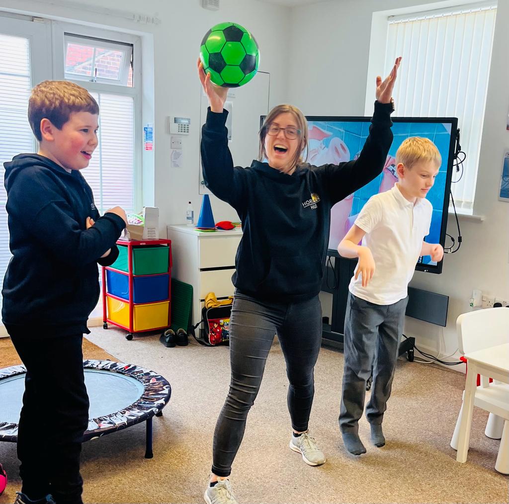 A facilitator is playing a game in a classroom with two younger children. The facilitator holds a ball above their head. Everyone is smiling and jumping.