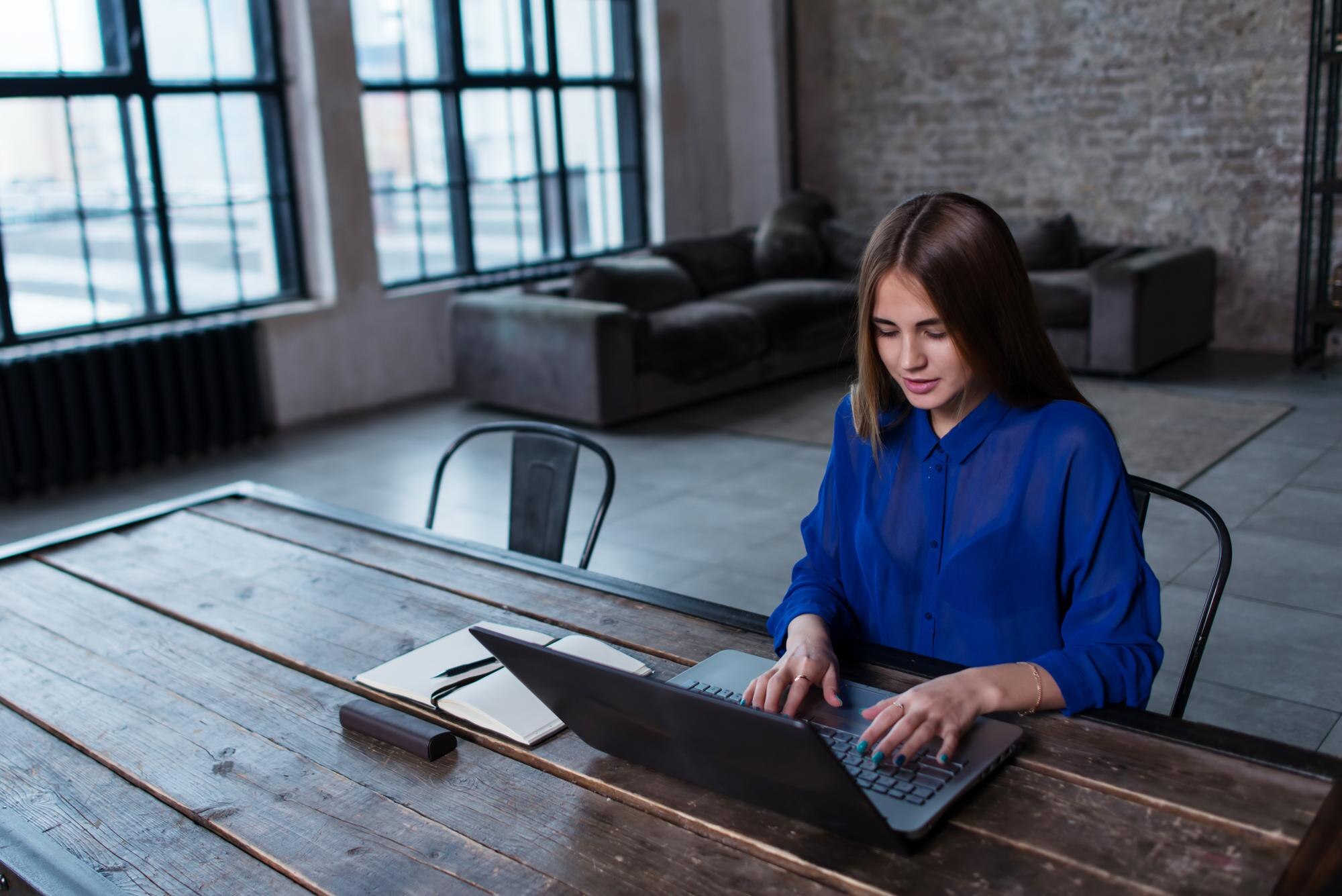 Person seated at a large wooden table using a laptop in a spacious office. An open notebook and pen lie beside the laptop, with large industrial-style windows, exposed brick walls, and seating visible in the background.