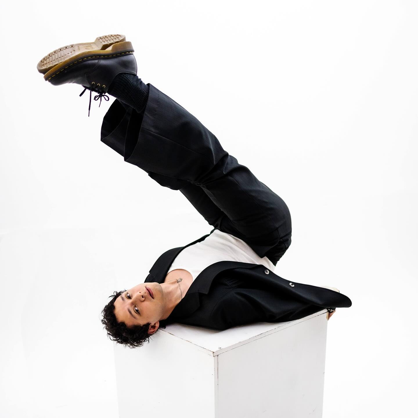 A person with curly hair dressed in a black suit and white shirt lying upside down with one leg propped high on a white cube, showcasing a tan-soled black boot against a white background.