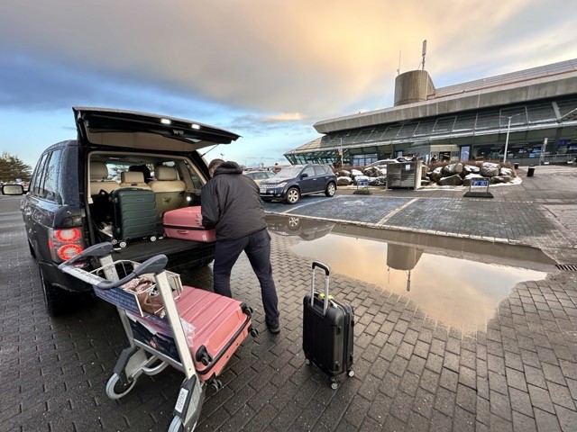 A traveler loading pink suitcases into the back of a vehicle outside an airport terminal, with reflections on wet pavement.