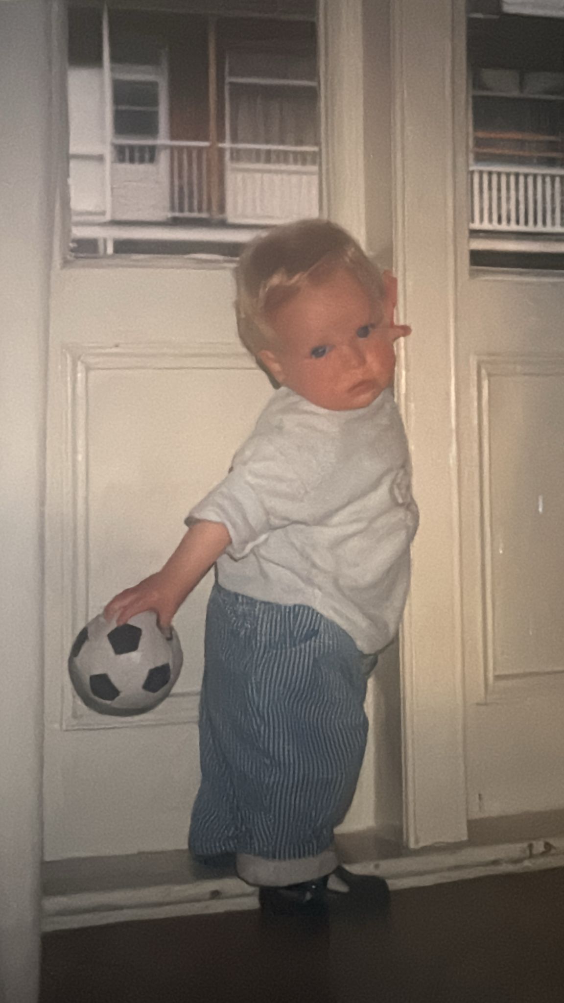 A groom in a dark suit carries a bride in a white dress across a sunny, grassy field. The couple shares a joyful, intimate moment with trees in the background.
