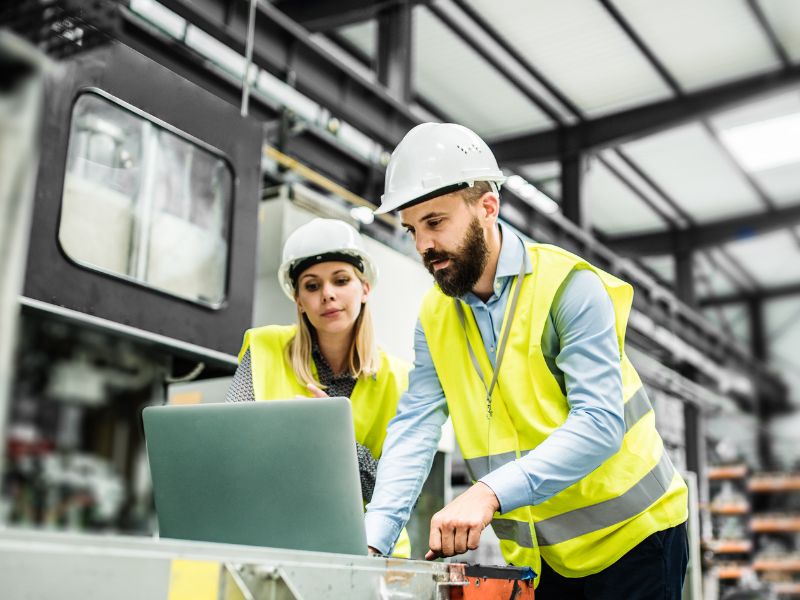 Two construction workers in hard hats and safety vests operate machinery in a bright industrial setting.