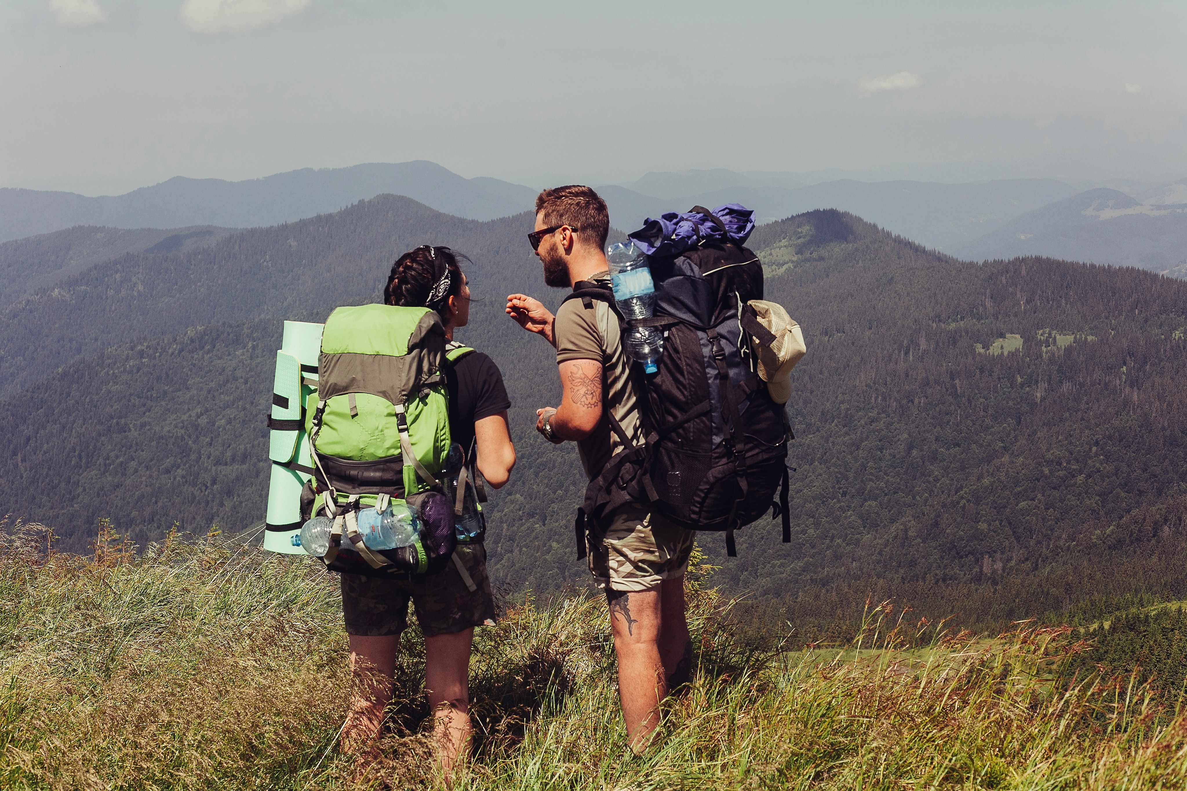 A man in an orange raincoat helps a woman in a yellow jacket navigate large rocks on the shore of a clear mountain lake.