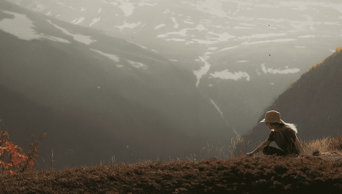 A landscape photo of misty mountains, covered in some snow. A person is sitting in the foreground,. wearing a sun hat. This person is the designer behind the website. The colours of the photo are warm and neutral.