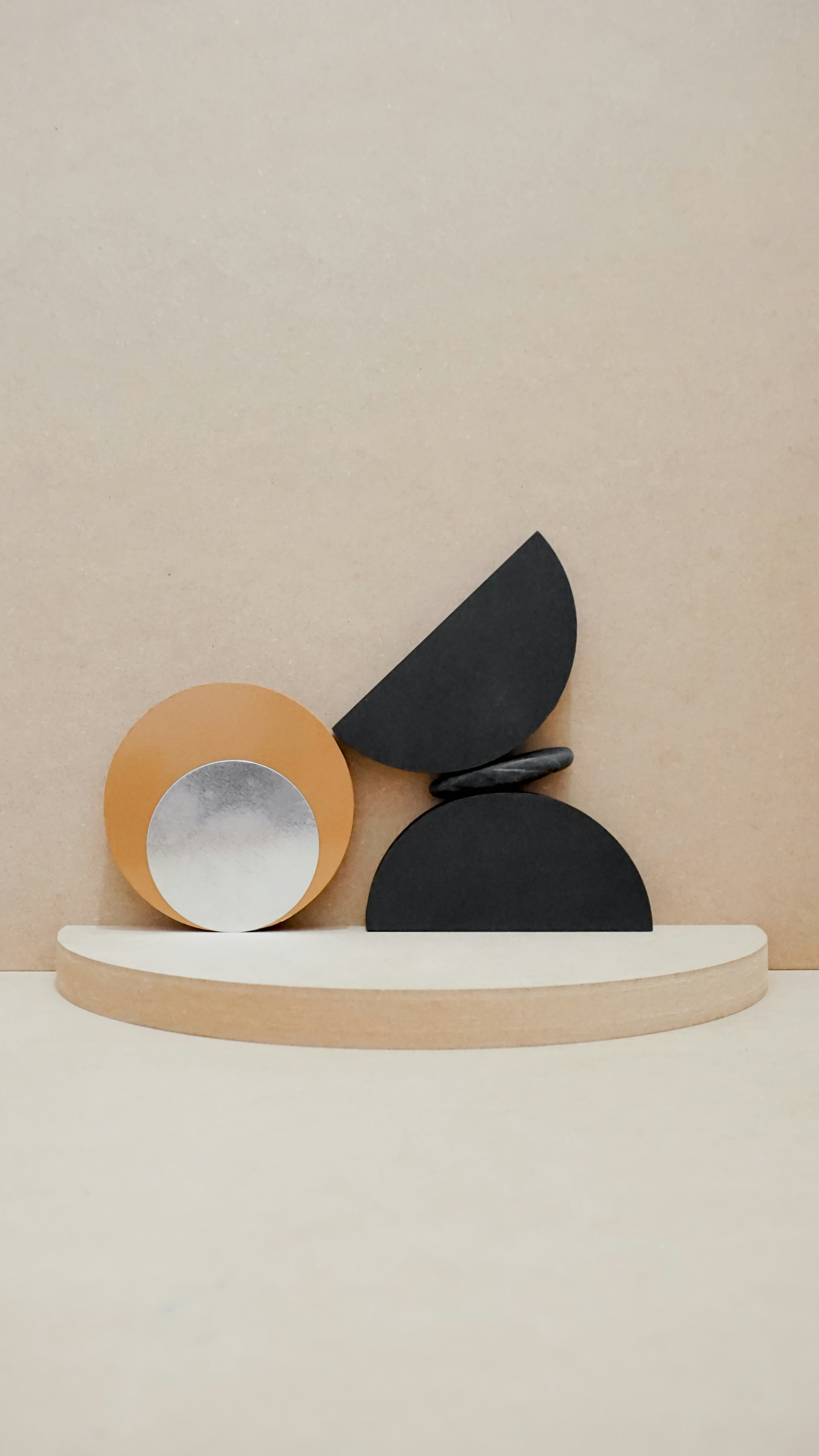 A minimalist abstract sculpture composed of geometric shapes on a light-colored semi-circular platform, featuring a large orange circle with a metallic silver disc inset, a black semi-circle at the base, a black quarter-circle balanced on top, and two small black oval shapes stacked between the black elements, set against a plain beige background.