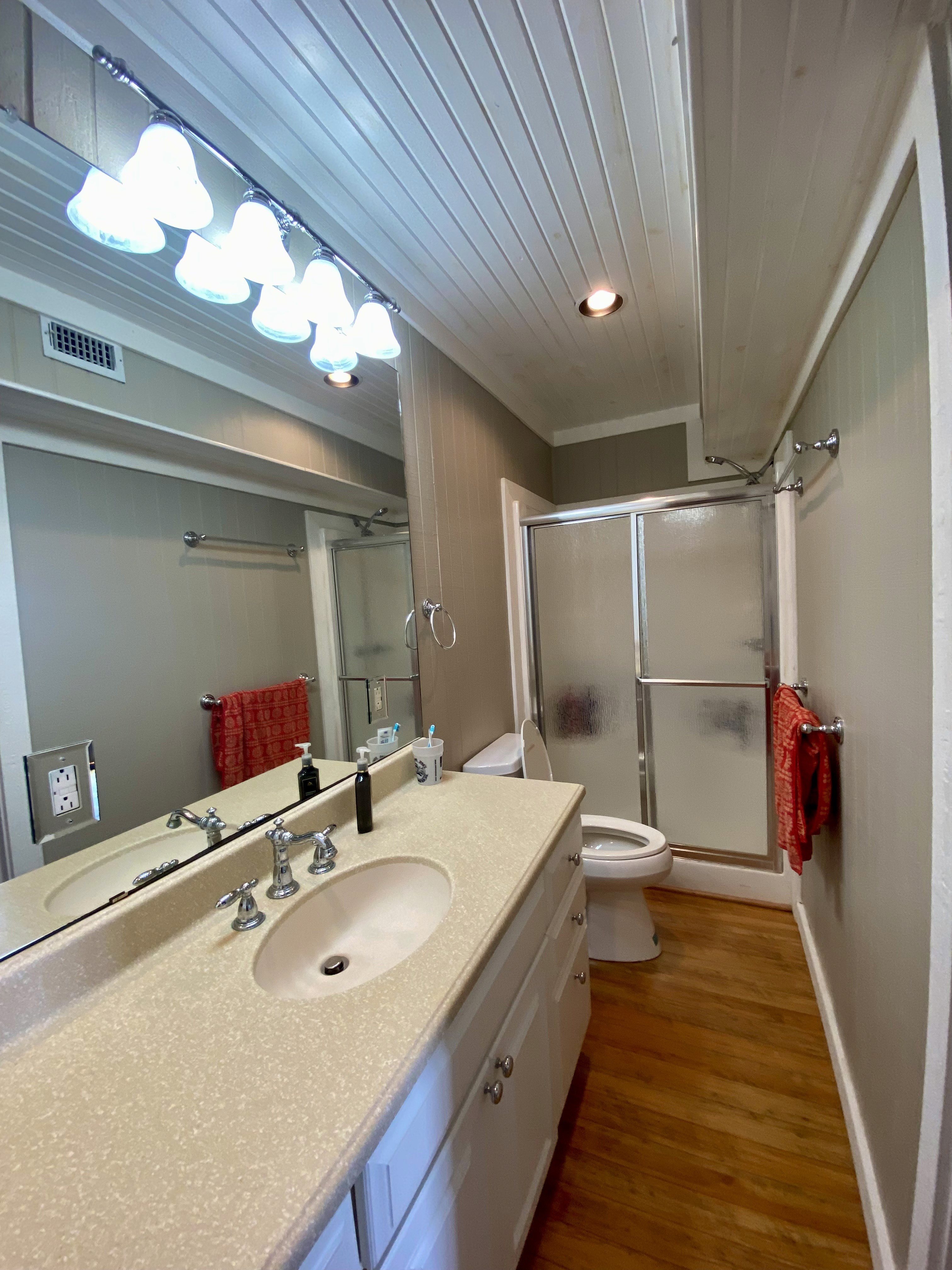 Bathroom with a vanity, large mirror, glass shower door, and wood flooring. Includes a towel rack with red towels.