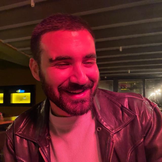 Person wearing a blue puffer jacket and red backpack standing in front of a light blue building on a city street.