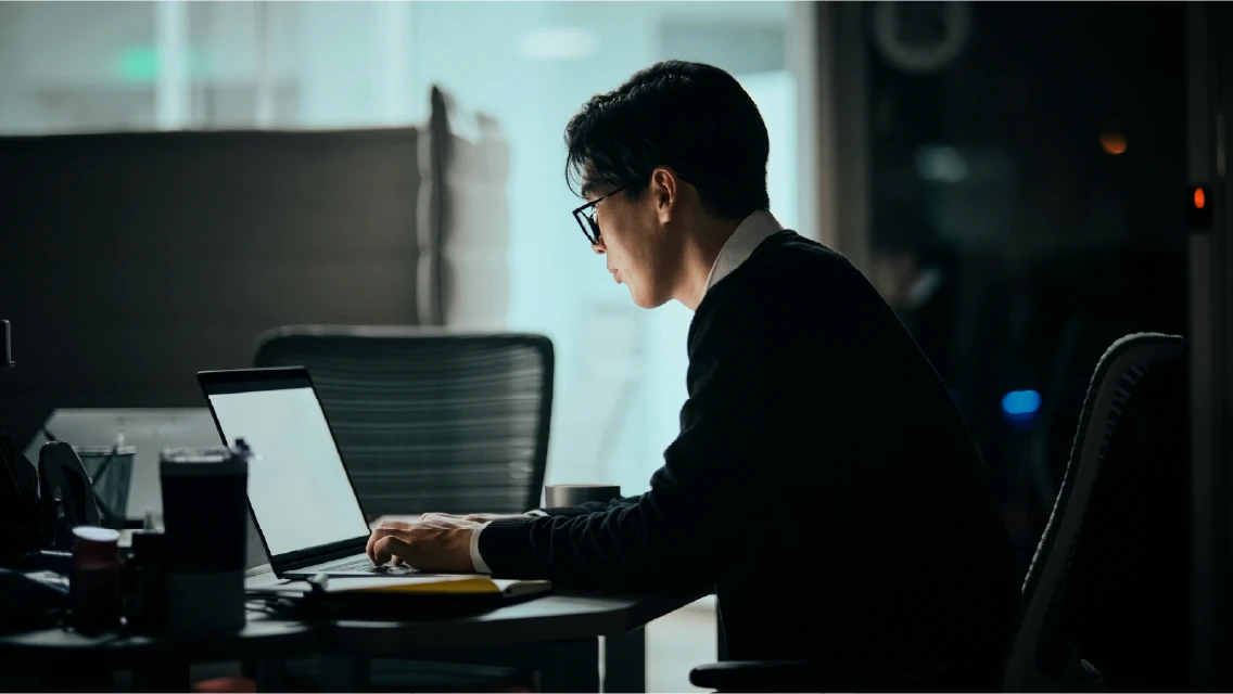 Silhouette of a remote worker at a laptop in a dim office, representing North Korean remote IT worker fraud schemes targeting distributed hiring.