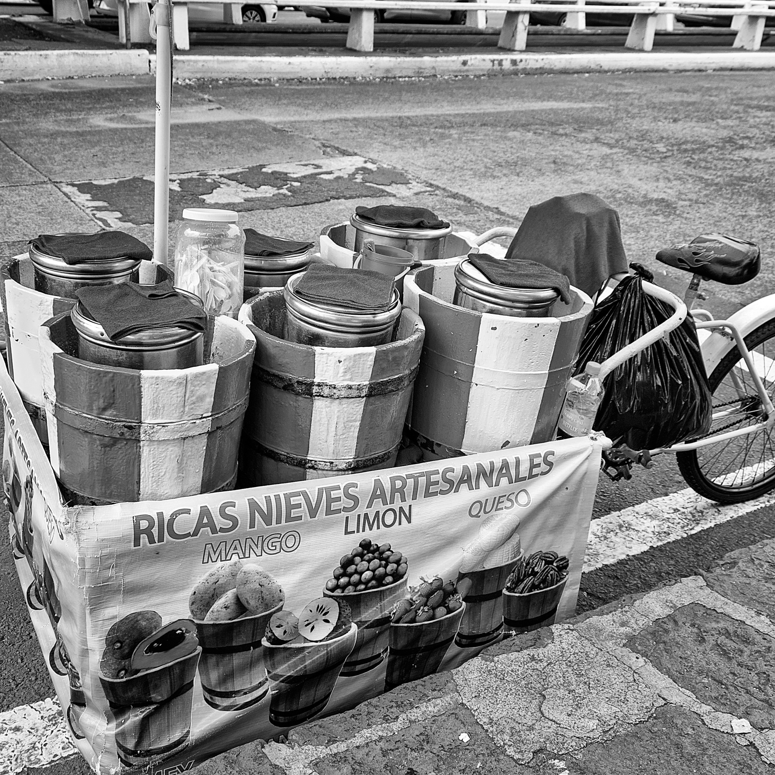 Black and white photograph of a bicycle cart selling artisanal ice cream, with wooden barrels labeled with flavors like mango, limón, and queso, parked on a city street.