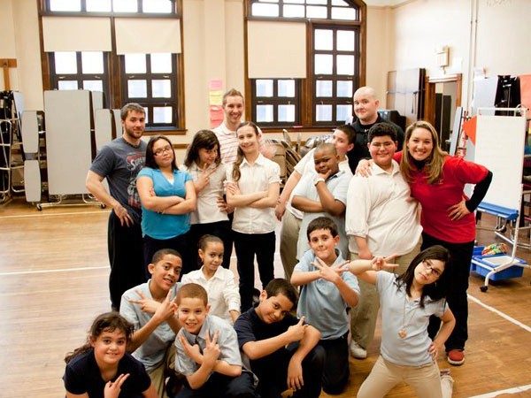 A group of approximately 20 people stands in a room with wooden flooring and large windows. The group is arranged in two rows, with some individuals standing in the back row and others in the front row. Many are making hand gestures, including peace signs and heart shapes. The group is dressed in a mix of casual and slightly more formal attire, with some wearing shirts and pants and others in t-shirts and shorts. The room has large windows that let in natural light, and a whiteboard is visible in the background. There are also some pieces of exercise equipment or storage units near the left side of the room.