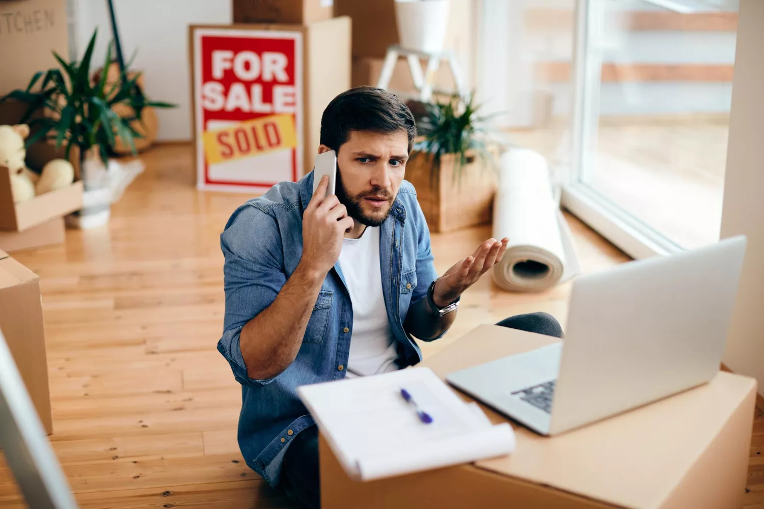 A man on his phone looks concerned while sitting among moving boxes and a laptop, symbolizing a seller facing unexpected e-commerce challenges.