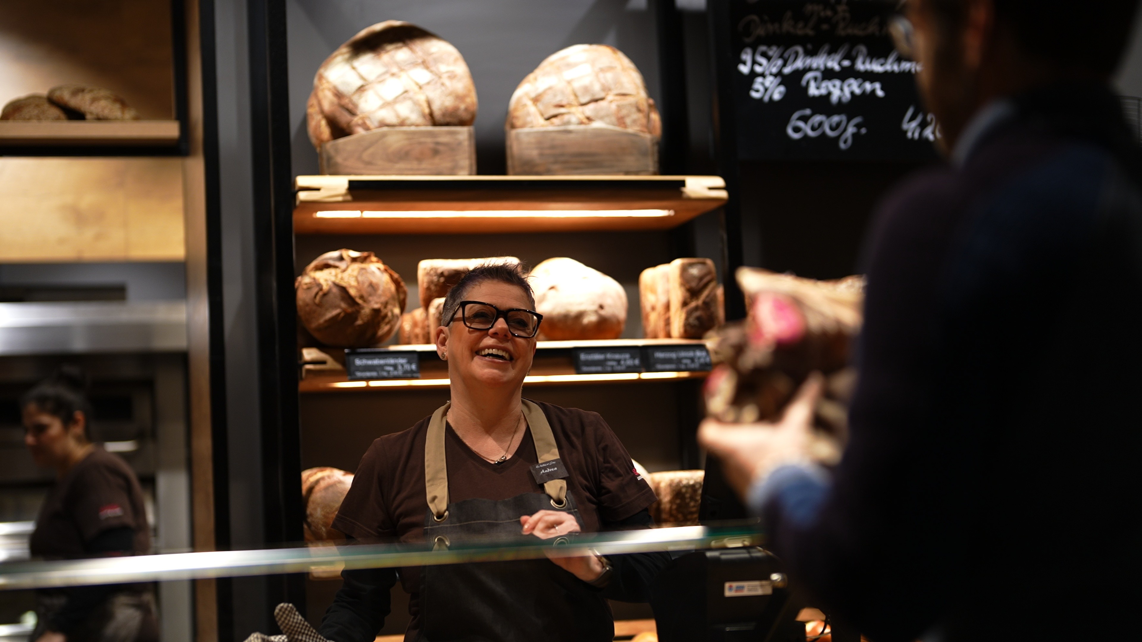 Mitarbeiterin lächelt einem Kunde hinter der Brot-Theke an.