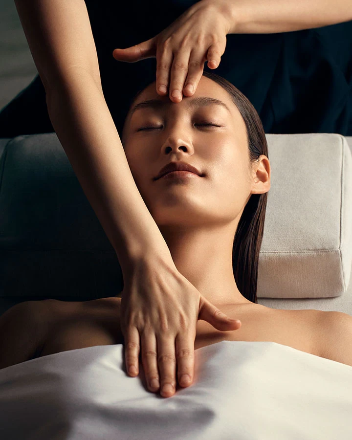 A woman receiving a facial massage, with one hand gently resting on her forehead and another hand applying pressure on her neck, all while she is peacefully lying down.