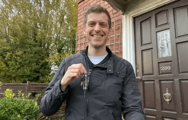 Man in grey jacket smiling and holding up house keys in front of a brick house entrance