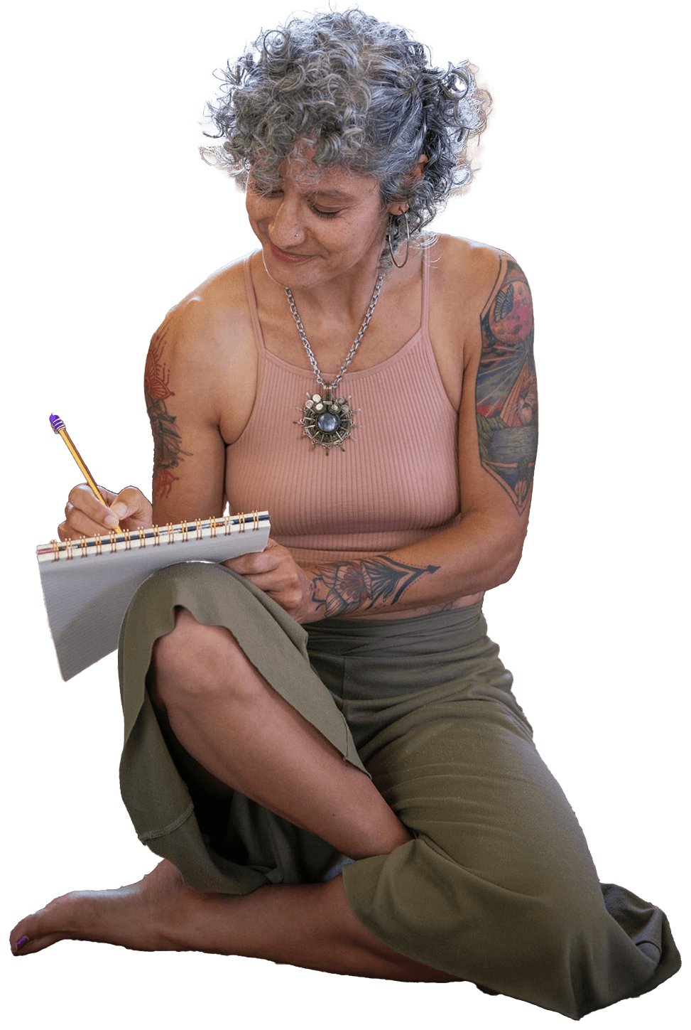 A woman with short gray hair and tattoos sits on the floor, writing in a notebook while wearing loose pants.