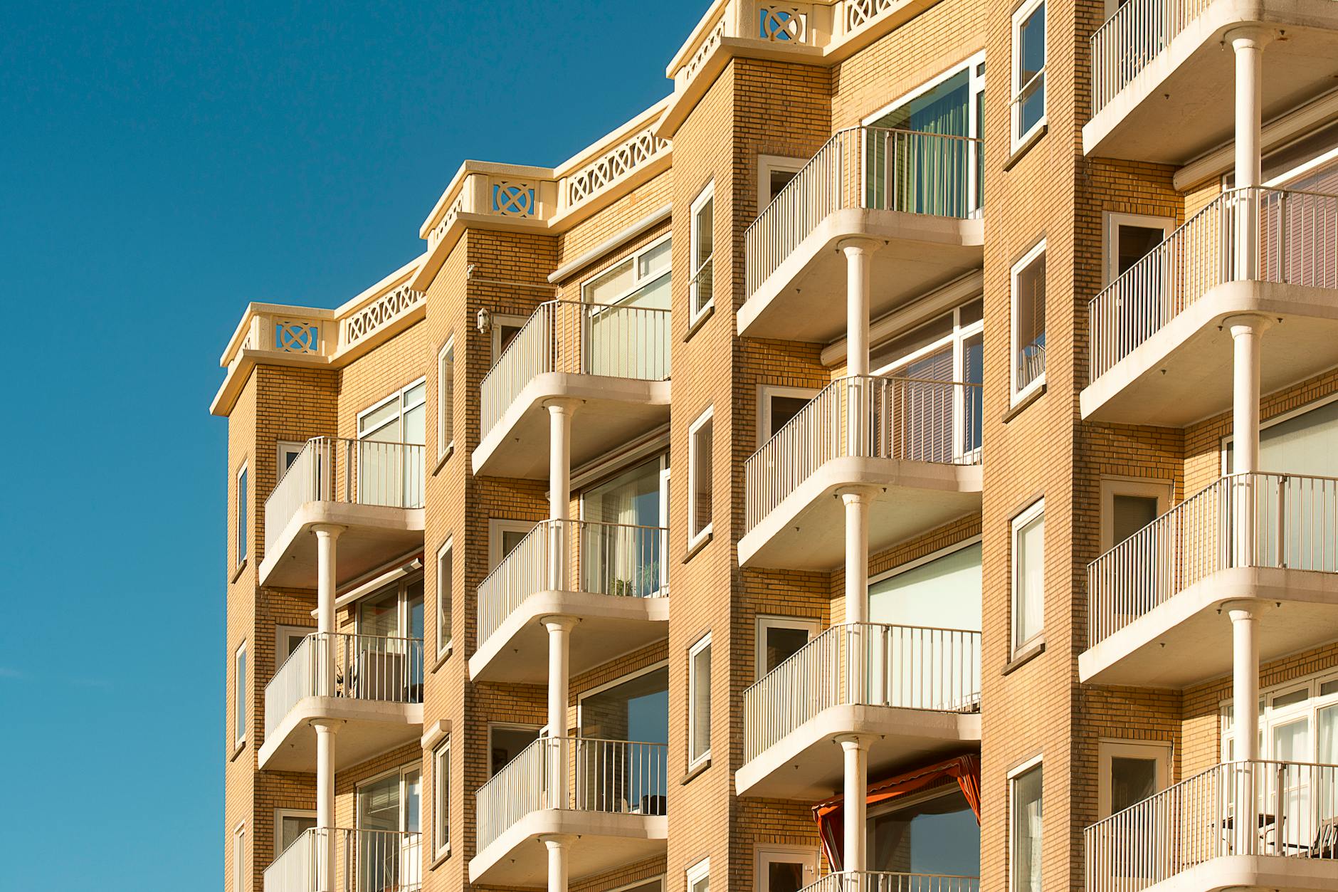 Facade of a sandy brick residential apartment block with white balconies against a clear blue sky