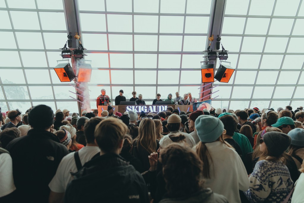 Crowded indoor concert with DJs at ski event, large windows, festive atmosphere.