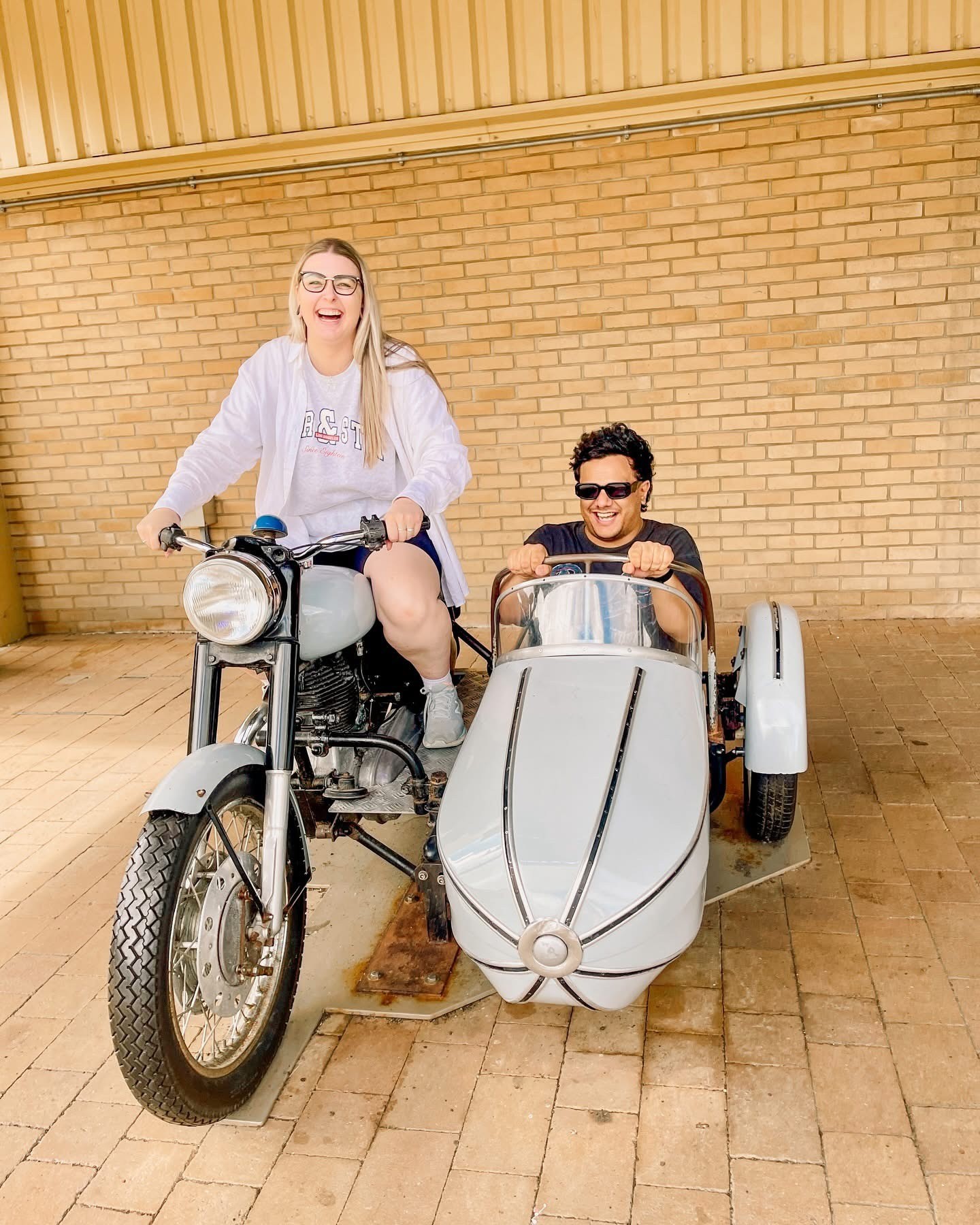 A woman with long blonde hair, in a brown top and jeans, walks through a sunlit forest, holding someone's hand. The scene is warm and inviting.