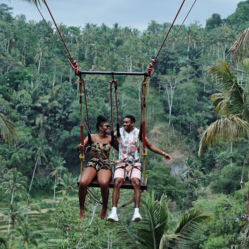 兩個人坐在高高懸掛的秋千上，俯瞰茂密的綠色森林地形和繁茂的棕櫚樹，面帶微笑，手牽著手。