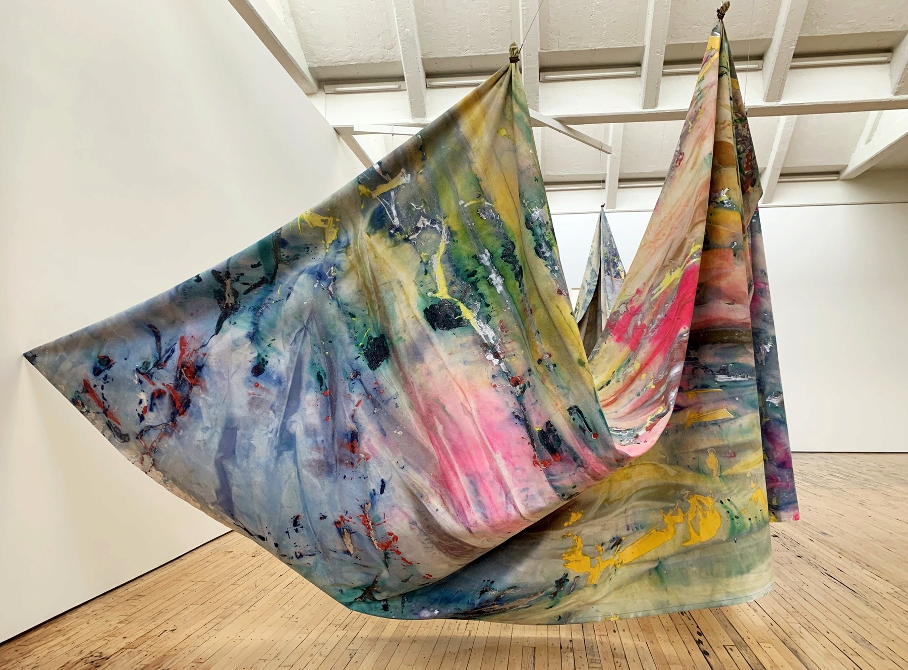 Large, draped fabric panels covered in vibrant paint splatters suspended in a white gallery space