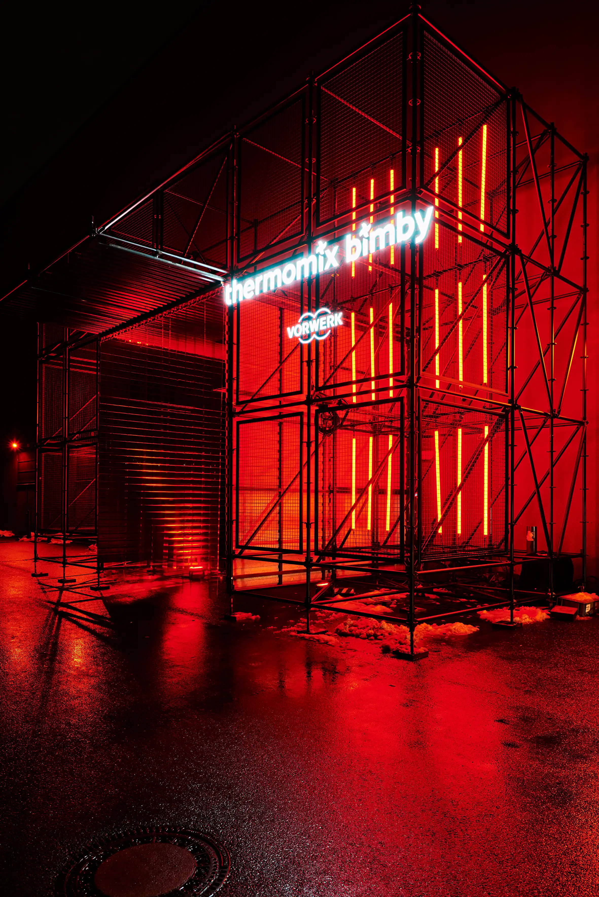 Scaffold entrance portal with neon Thermomix Bimby sign and red light strips at night.