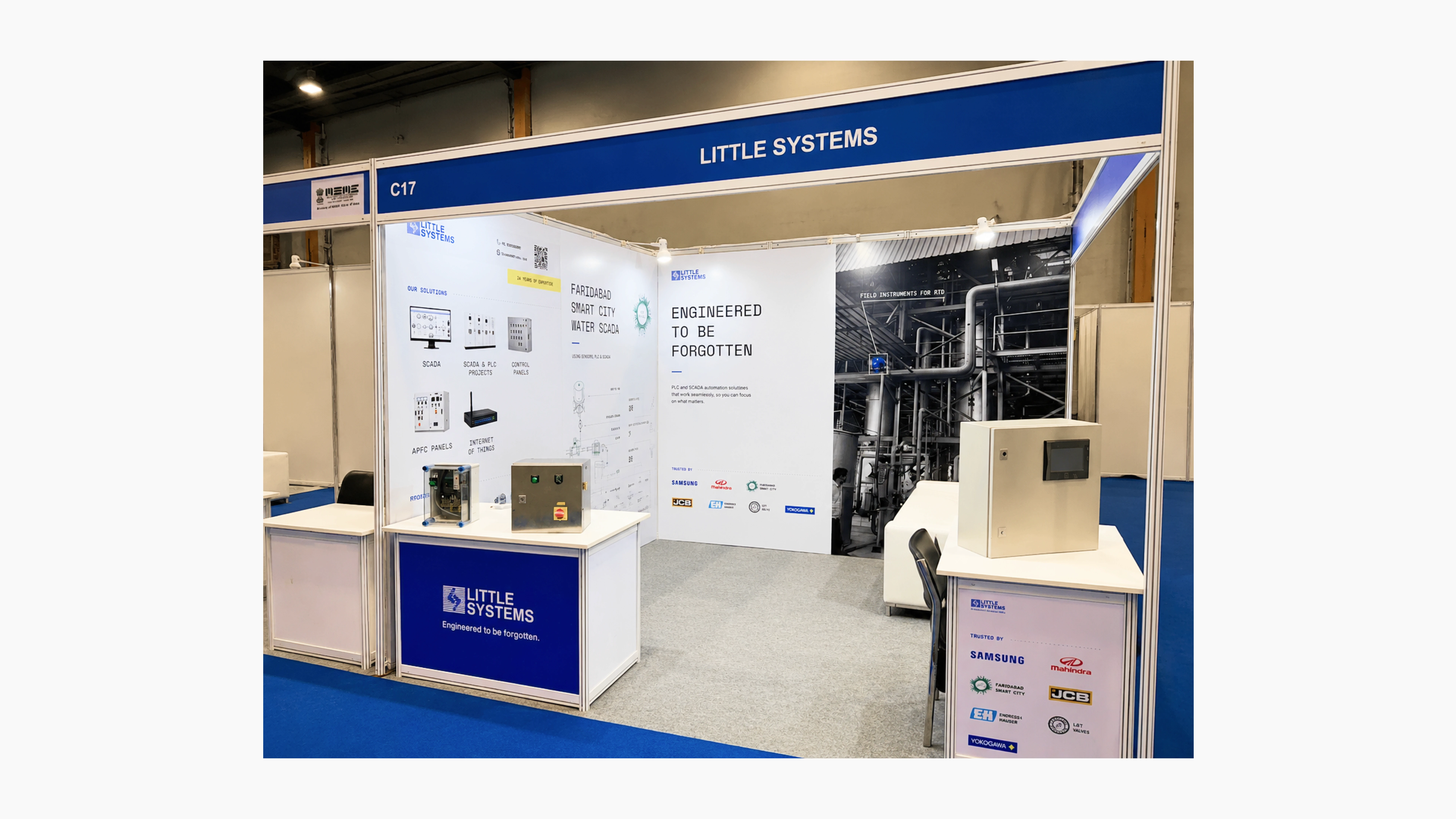 Wide view of the Little Systems exhibition booth at an industrial trade show, featuring blue-and-white branding, automation equipment displays, technical posters, industrial photography, and demonstration control panels.