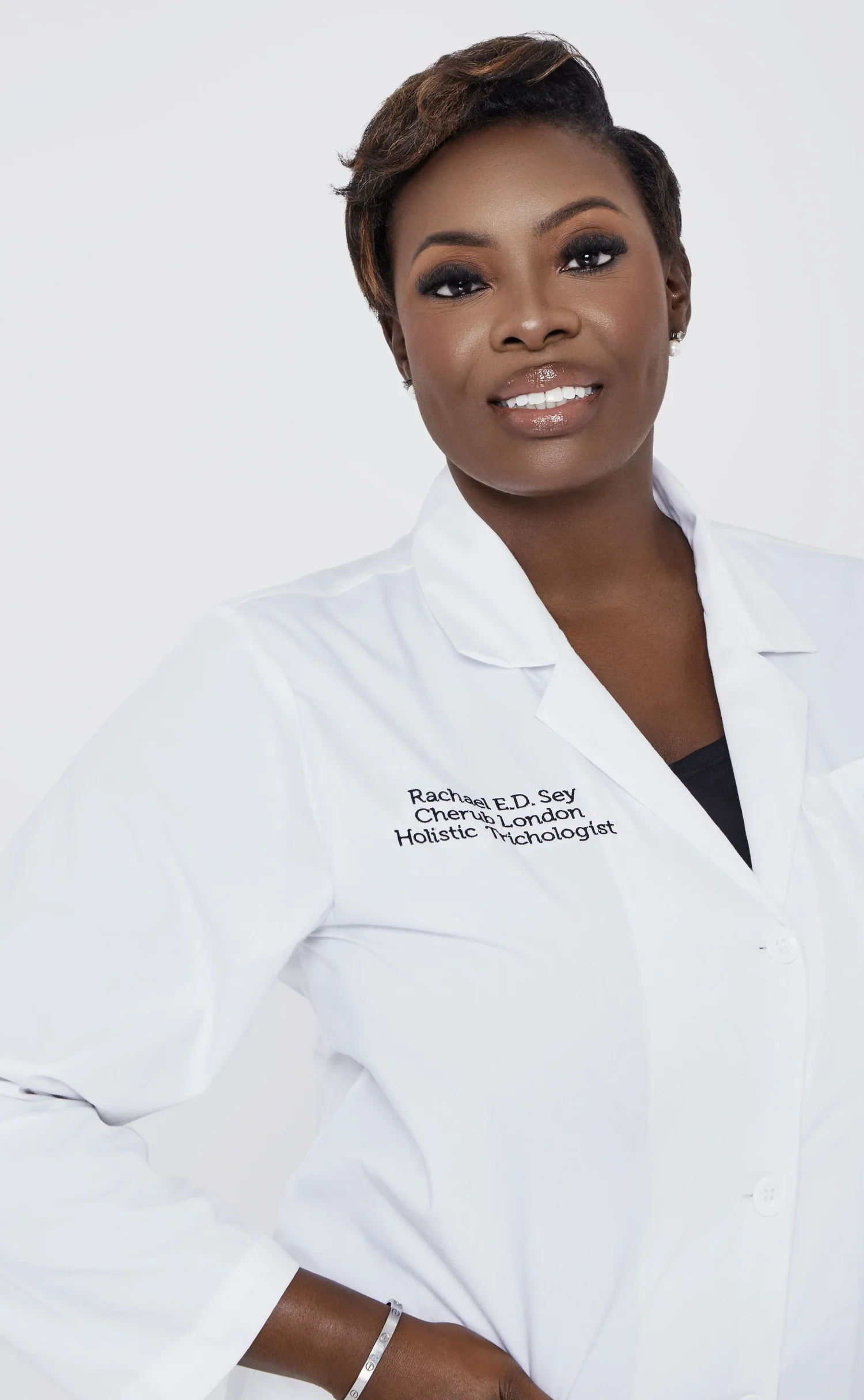 Smiling male doctor in a white lab coat and scrubs, standing with arms crossed in a warm, modern clinic.