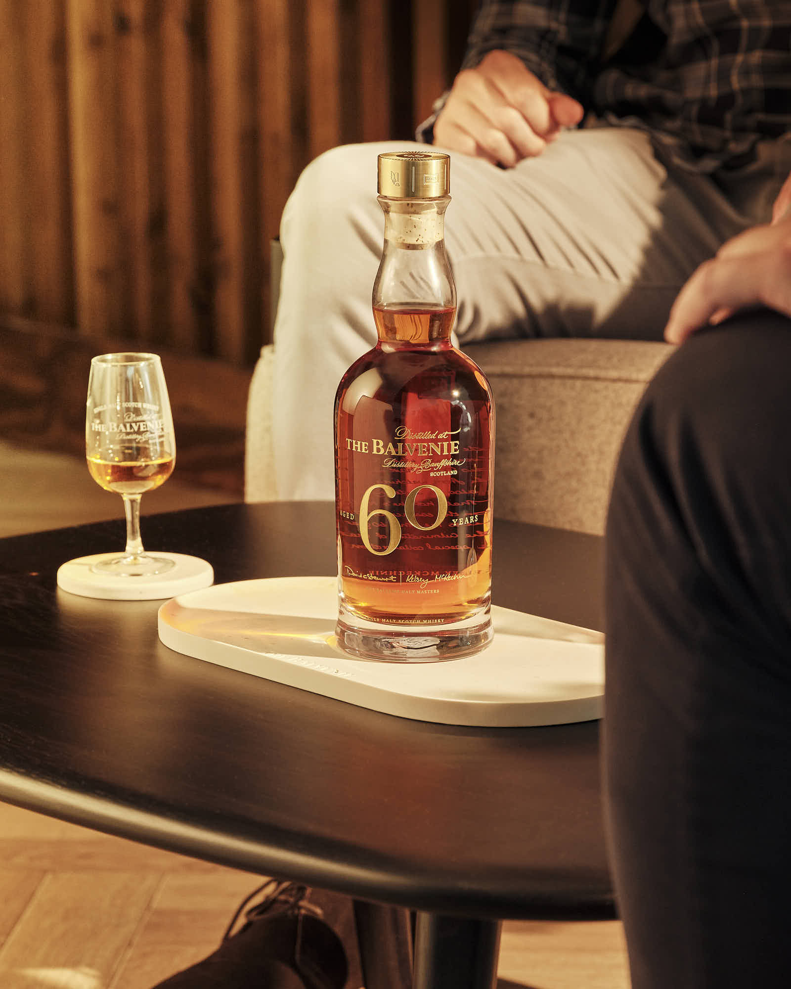 A whiskey bottle and a glass sit on a table, with a cozy sofa and person lounging in the background.