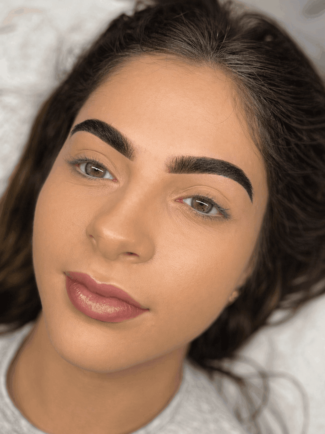 Young woman with full, dark eyebrows lies on white surface, appearing relaxed and content. Her skin is smooth, and she has a slight, serene smile.