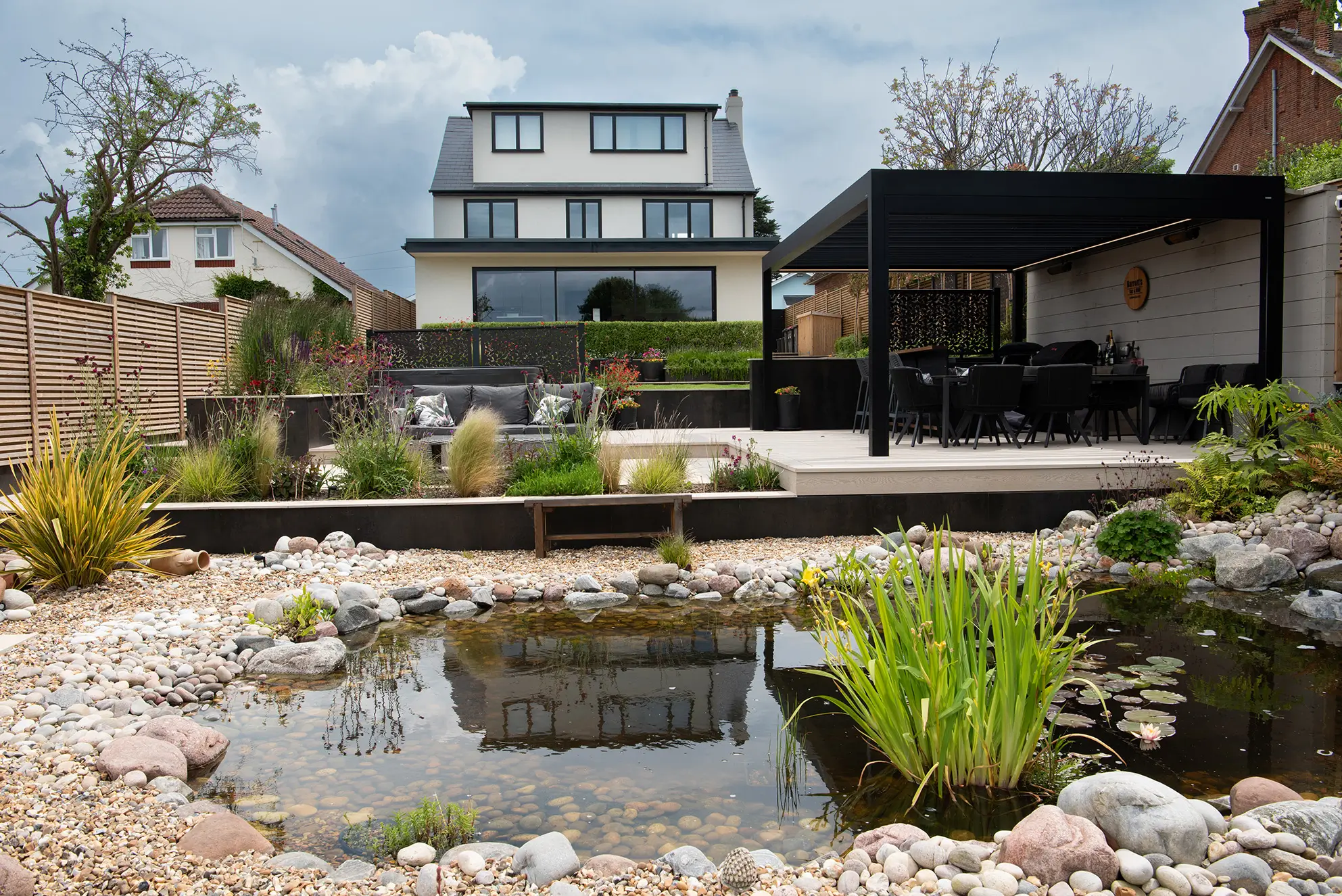 A landscaped garden with a pond, plants, and a modern building in the background under a partly cloudy sky.