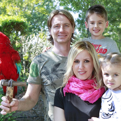 一家四口站在户外，一只红色鹦鹉栖息在一个男人的手臂上。背景中是树木和绿色植物。