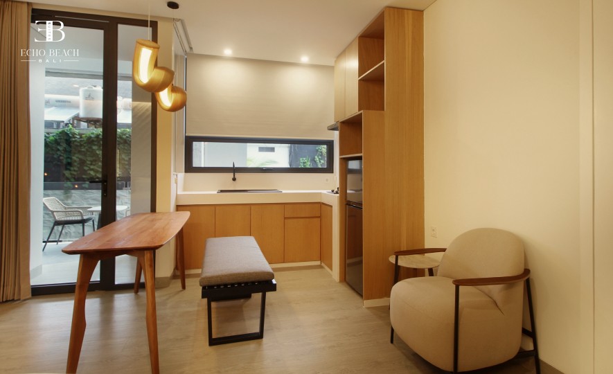 Dining and kitchenette area in an Echo Beach Suite, Canggu, Bali, featuring a wooden table, a beige armchair, a kitchenette with a window above the sink, and a unique pendant light.