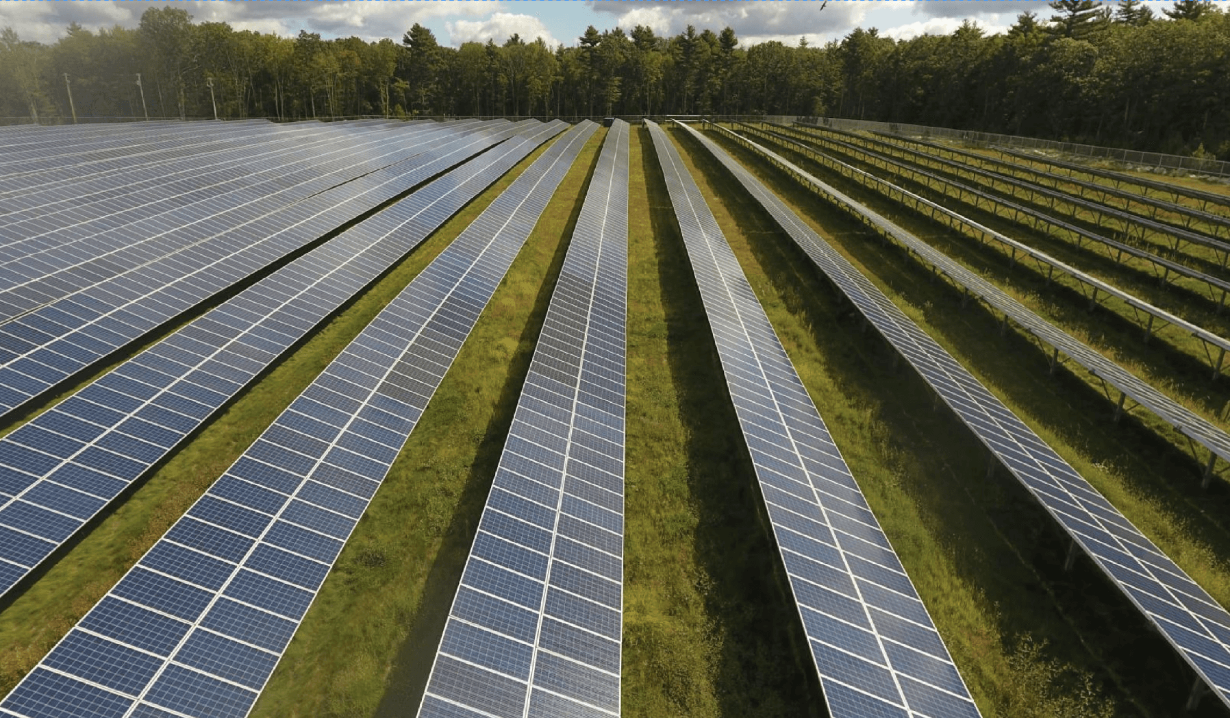 Aerial view of a solar farm with rows of solar panels surrounded by greenery and cloudy skies.