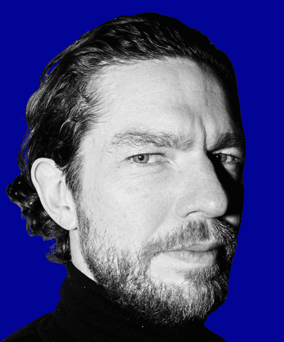 A young person with curly hair smiles at the camera while sitting in front of a laptop against a dark background.